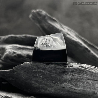 An image of the Kord Himalayan Mounts Artisan Keycap. This artisan keycap made of resin is portrayed in an earthy background surrounded by dark contrasting rocks to symbolize mountains. This artisan resin keycap is also shown with mist in the background emulating a cool mysterious night